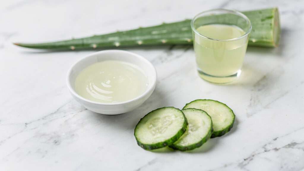 DIY aloe vera and cucumber face mask for sensitive skin — small white bowl of aloe vera gel, fresh cucumber slices and a glass of cucumber juice on white marble.