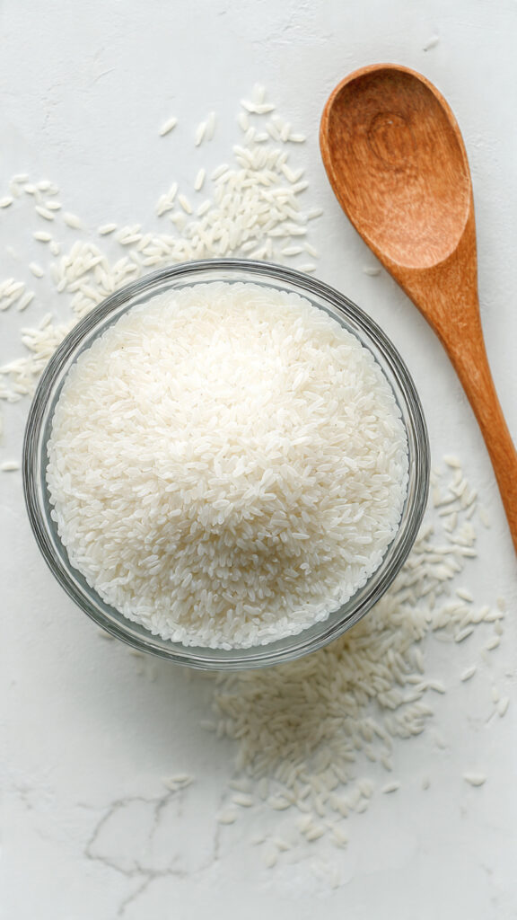 Plain milky rice water in a clear glass bowl on white marble with uncooked rice and wooden spoon